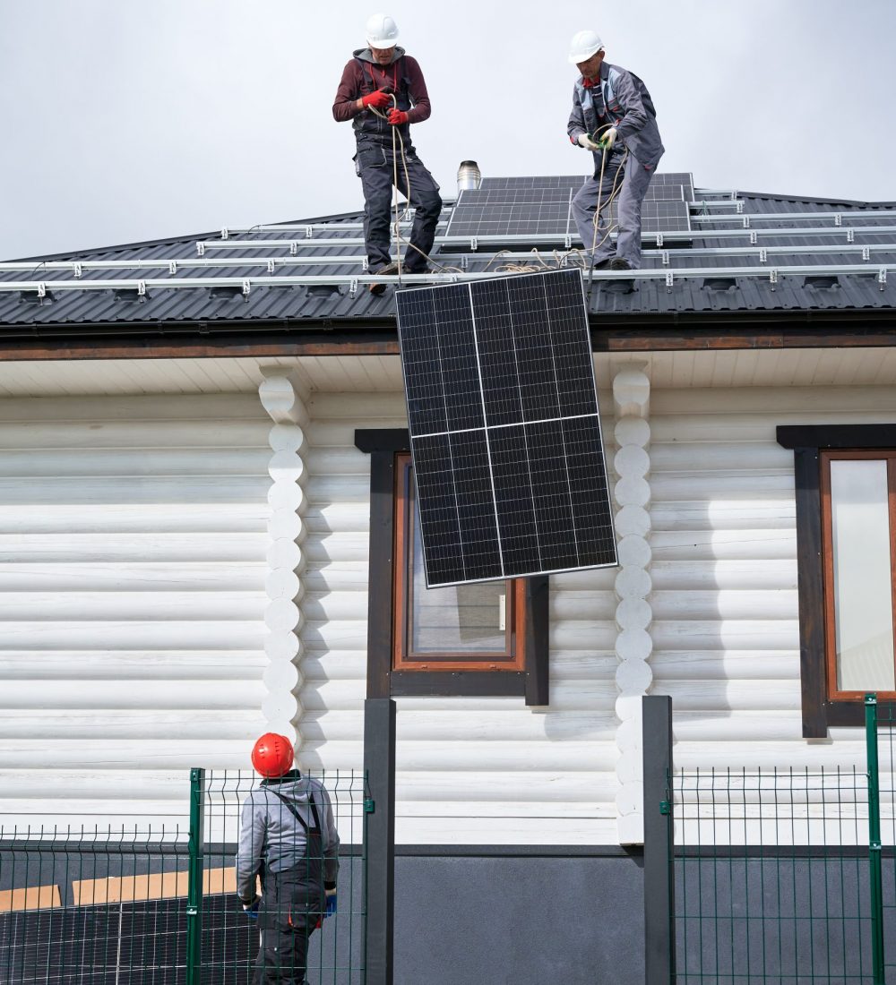 Men workers installing solar panel system on roof of house. Electricians in helmets lifting up photovoltaic solar module with help of ropes outdoors. Concept of alternative and renewable energy.